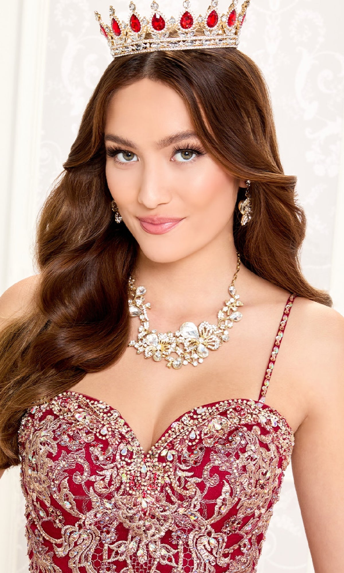A woman wearing the Quinceanera Dress PR30086 By Princesa, featuring a jeweled crown, ornate red and gold design, sheer cape, and statement necklace, poses smiling in front of a light patterned background.